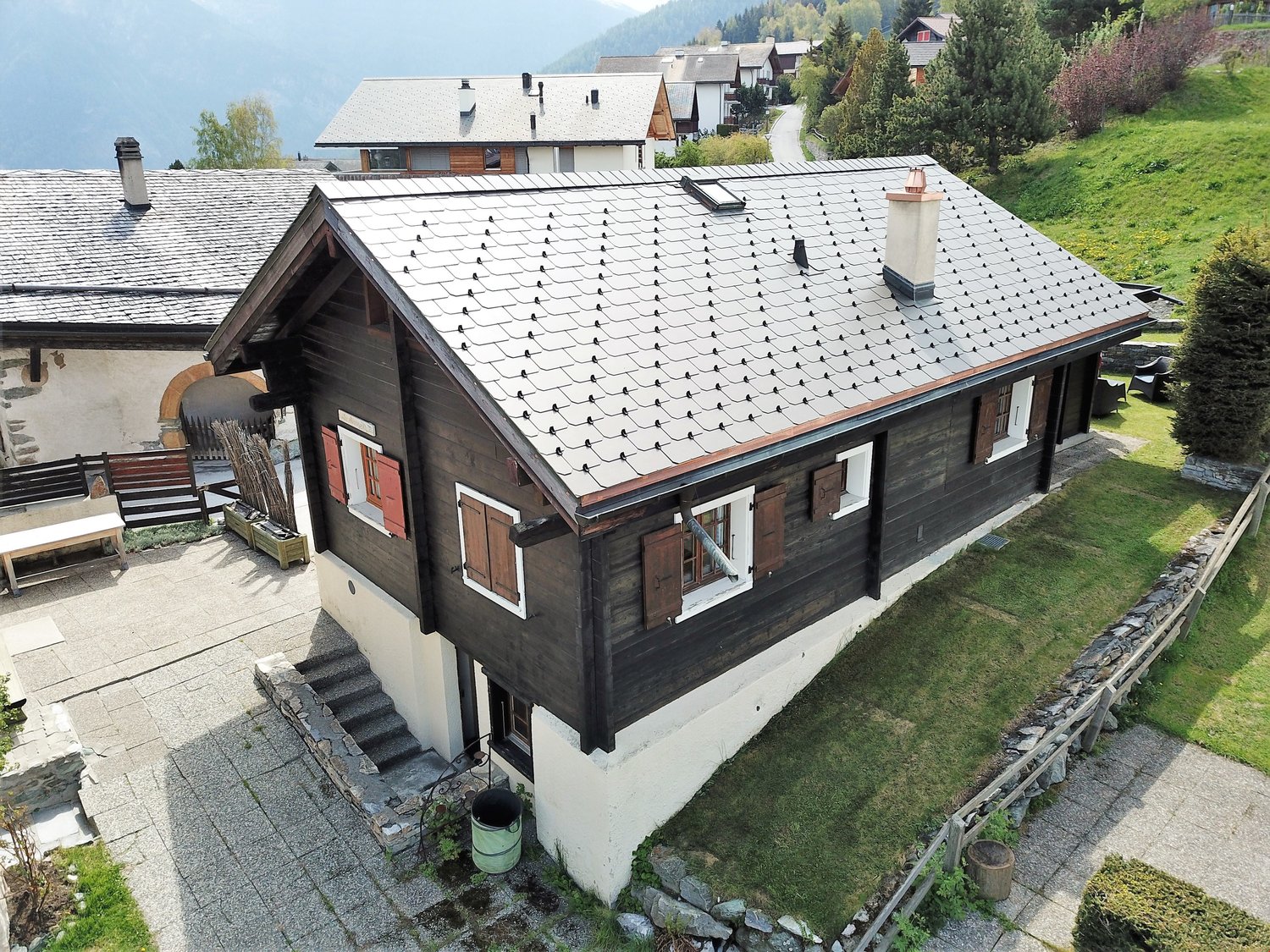 2 story wooden chalet-style house with a slate roof, surrounded by a grassy area and trees. The house has a balcony or terrace on the upper level.