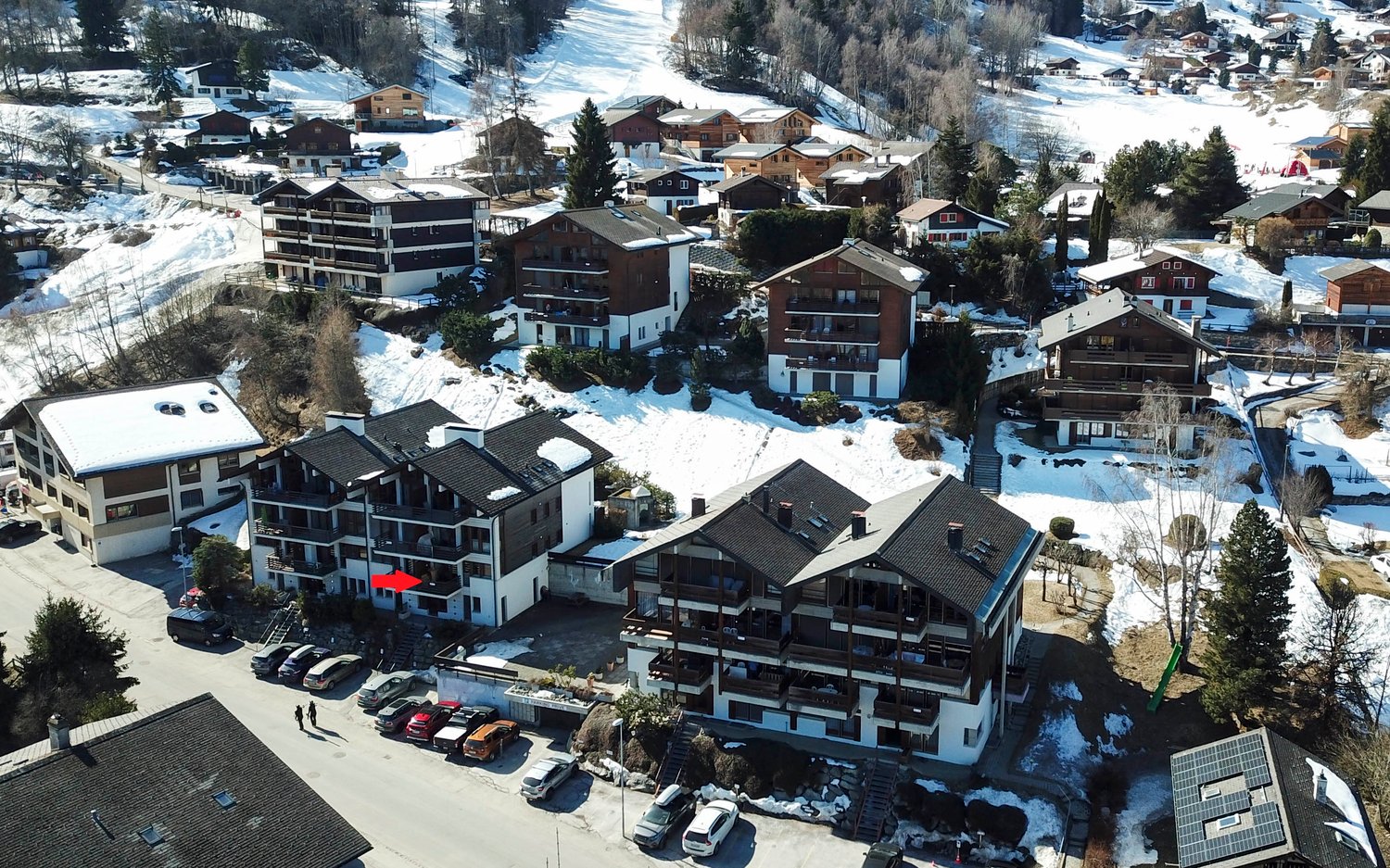 Aerial view of a snowy mountain village with multi-story apartment buildings, chalets, and parking spaces. The village is surrounded by snow-covered trees and slopes, indicating a ski resort or alpine setting.