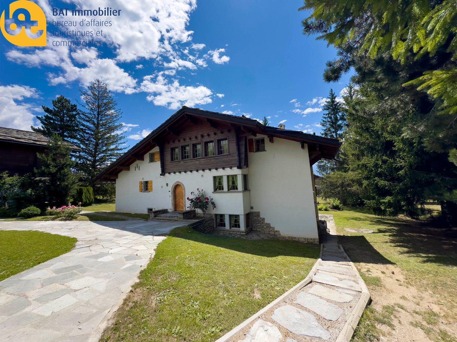 2-story chalet-style house with wooden beams, white walls, and large windows. The house is surrounded by a lush green lawn and pine trees, with a stone pathway leading to the entrance. There appears to be a balcony or terrace on the upper level.