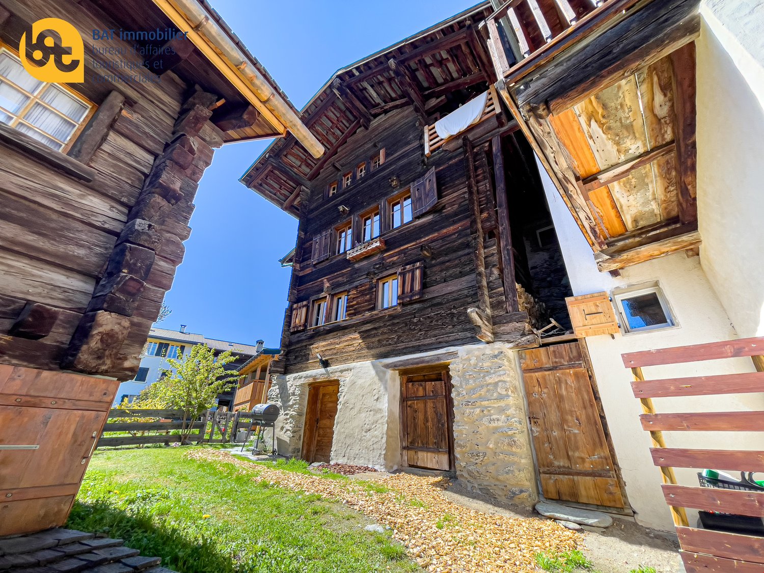 Two-story wooden building with stone foundation, wooden balconies, and large windows. The building has a rustic, traditional alpine style architecture. There is a grassy area in the foreground with some gravel and a wooden bench.