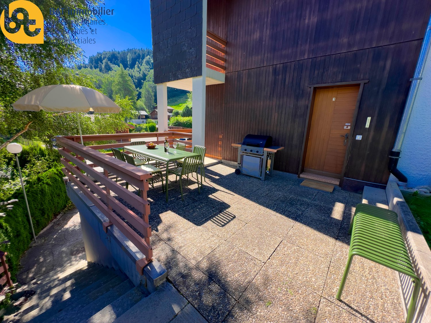 Wooden exterior, covered patio with dining table and chairs, barbecue grill, parking area, surrounded by greenery and mountains