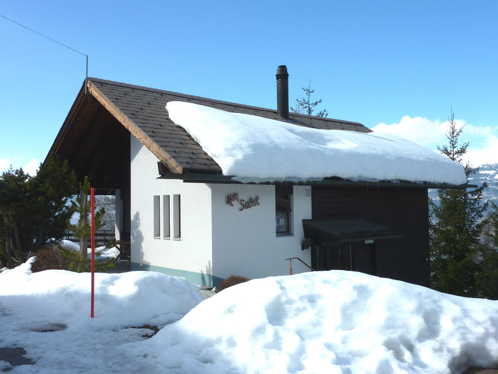 2-story wooden chalet-style house with a slate roof, surrounded by snow-covered landscape, with a chimney and a balcony or terrace visible.