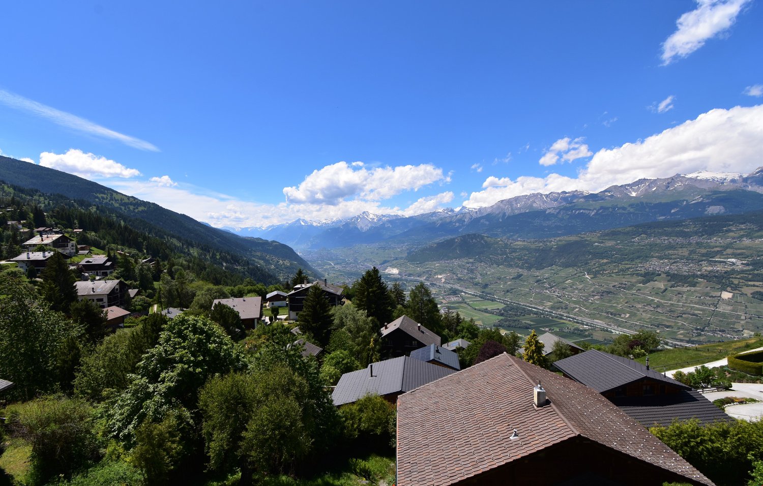 The image shows a scenic mountain landscape with a valley below. There are several houses and buildings nestled among the trees on the hillside, with snow-capped peaks visible in the distance. The sky is clear and blue, with some fluffy white clouds. The