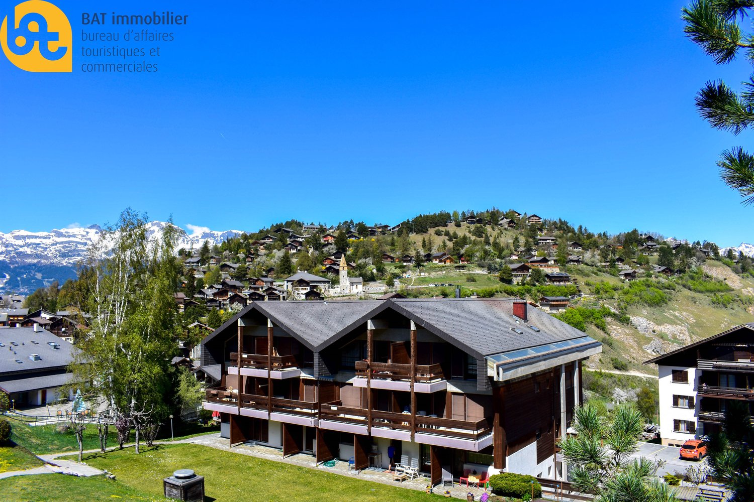 Multi-story wooden chalet-style building with balconies, surrounded by a grassy area and pine trees, with a village and mountains in the background.