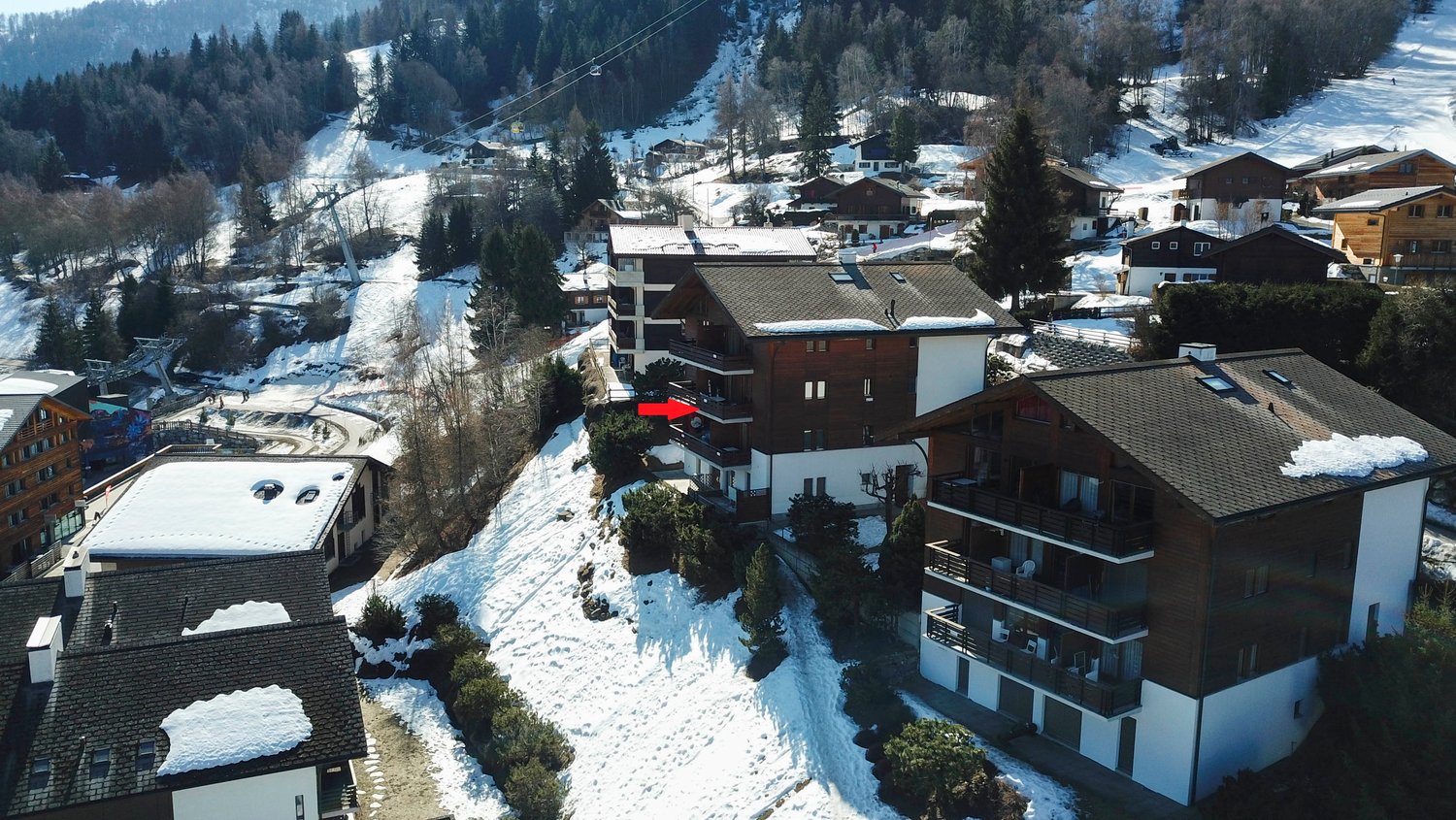 Aerial view of a ski resort town with snow-covered buildings, ski slopes, and cable cars in the background. The image shows a residential area with apartment buildings and houses, as well as some commercial buildings.