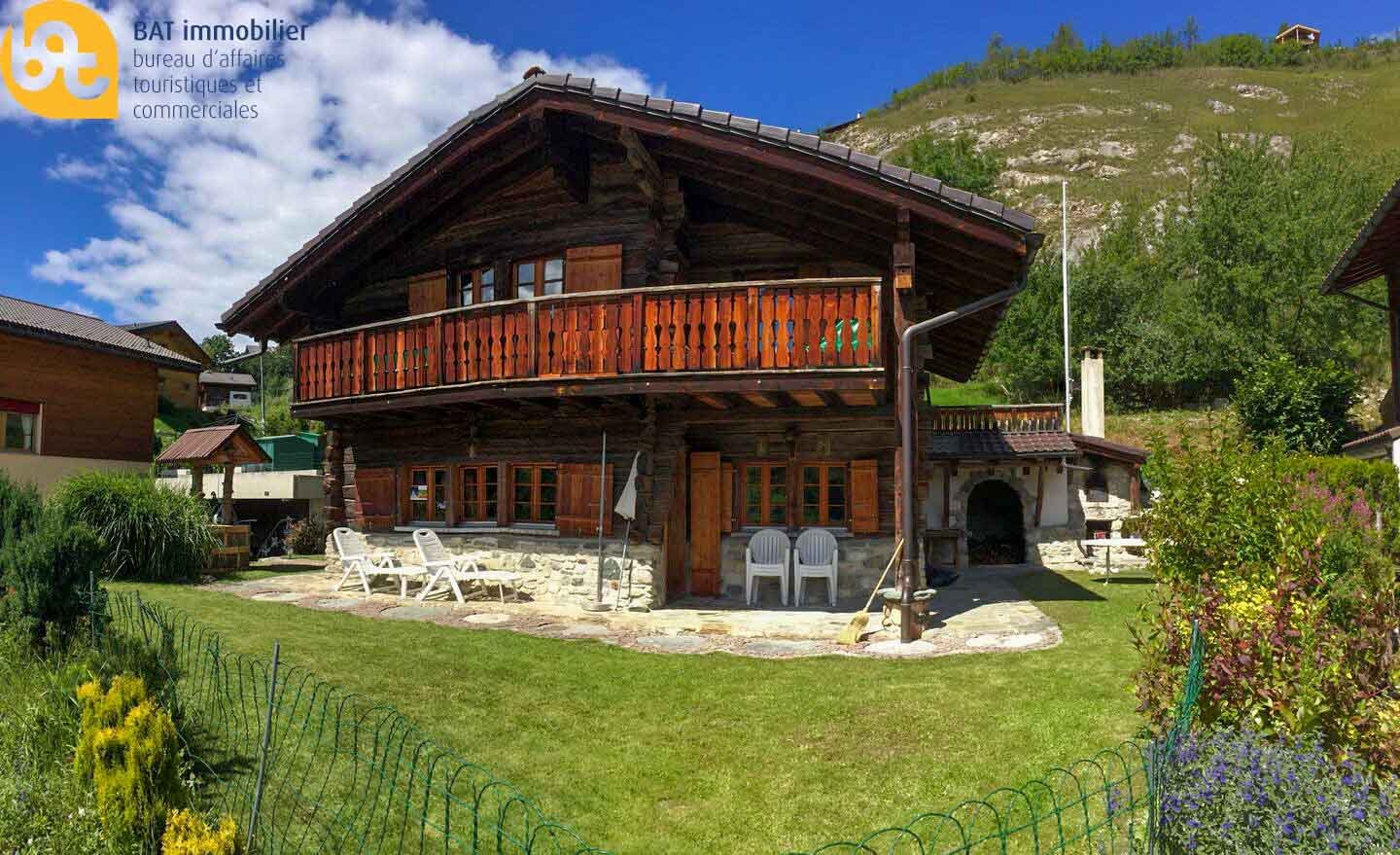 2-story wooden chalet with a balcony, surrounded by a grassy yard and mountains in the background. The chalet has a stone fireplace and outdoor seating area.