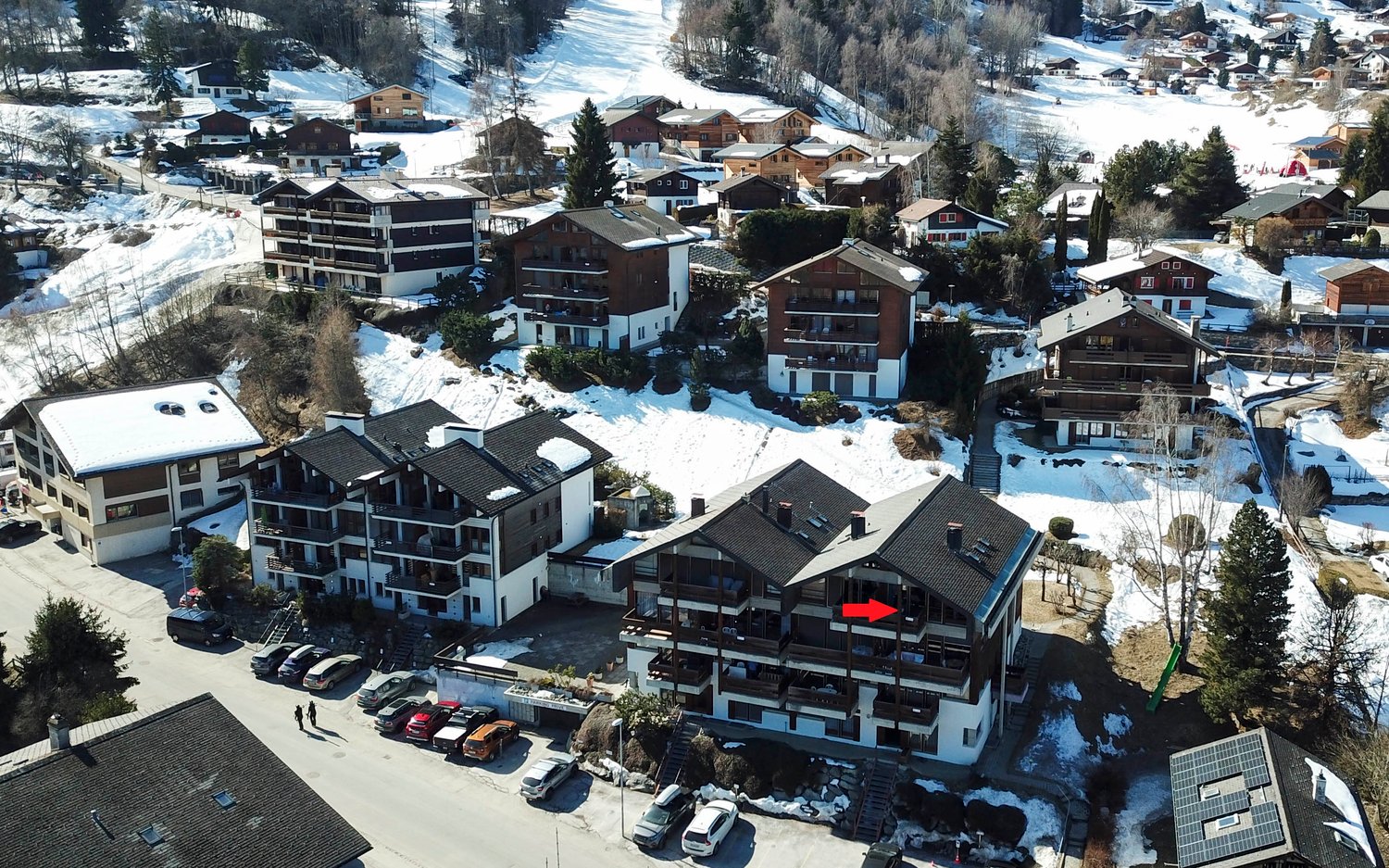Aerial view of a ski resort town with multiple apartment buildings, chalets, and parking areas surrounded by snow-covered mountains and trees.
