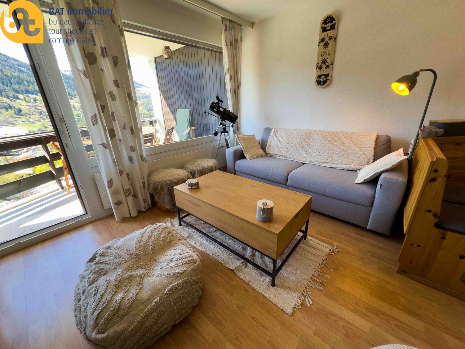 Cozy living room with large windows, gray sofa, wooden coffee table, and a telescope on a tripod. The room has a balcony or terrace with a scenic mountain view.