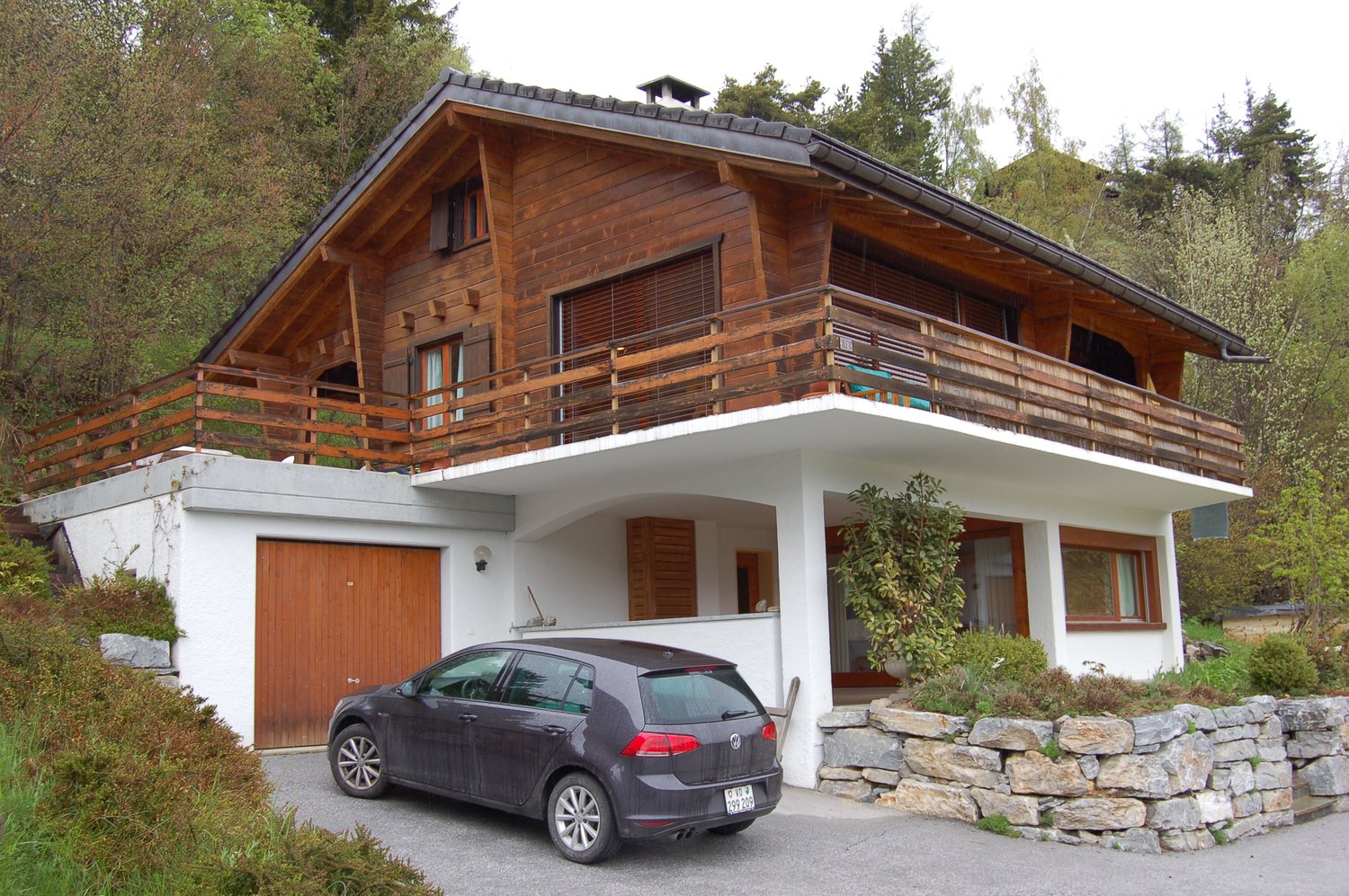 2-story wooden chalet-style house with a garage, balcony, and stone foundation. The house has a slanted roof and large windows. There is a car parked in the driveway in front of the house.