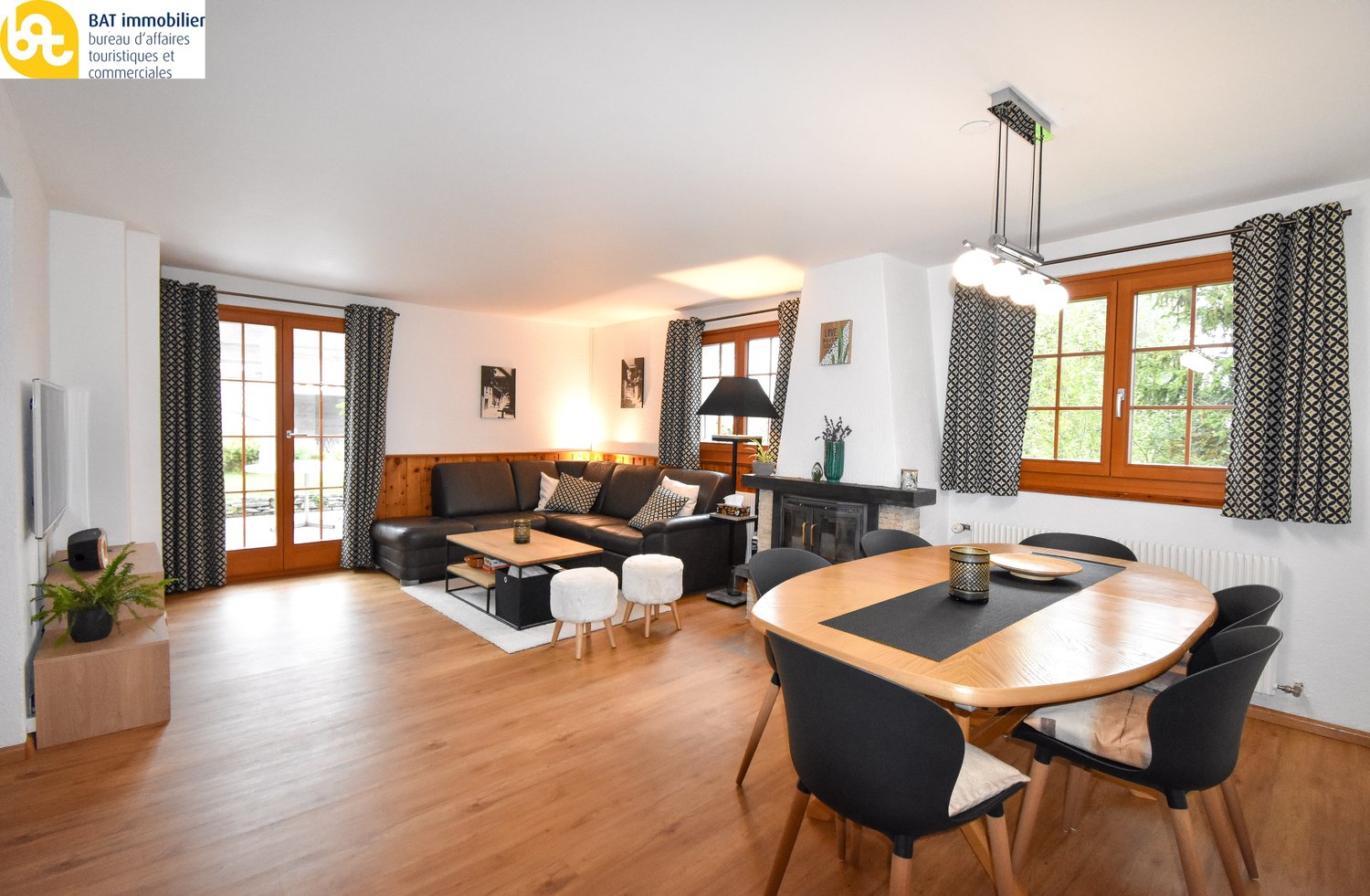 Spacious open-plan living area with hardwood floors, white walls, large windows, black leather sofa, wooden dining table and chairs, fireplace, and decorative lighting fixtures