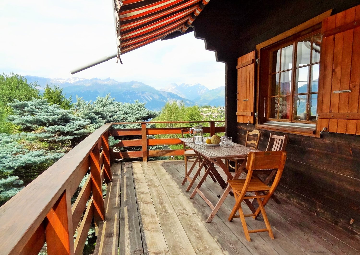 Wooden patio with a table and chairs, surrounded by pine trees and mountains in the background. The patio has a striped awning overhead providing shade.