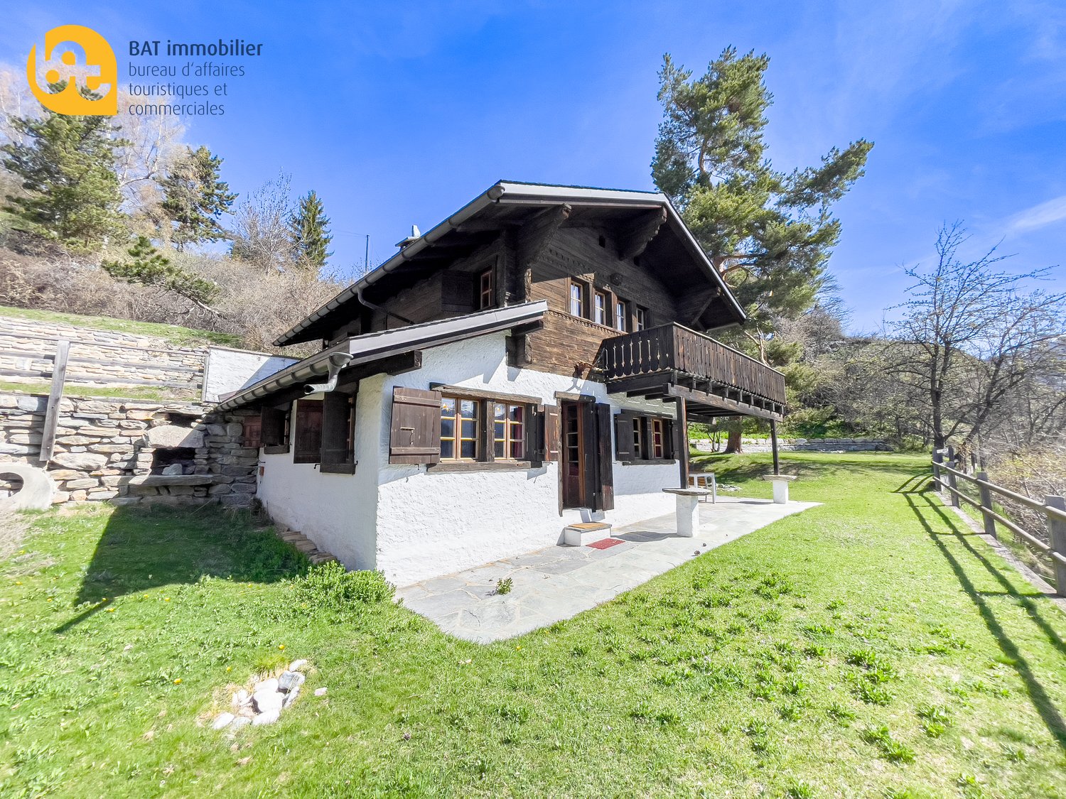 2-story chalet-style house with wooden exterior, stone walls, large balcony, surrounded by pine trees and green lawn