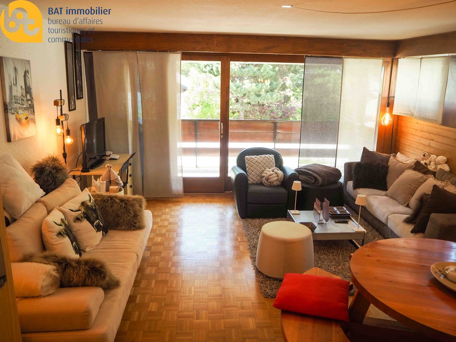 Spacious living room with large windows overlooking a balcony, hardwood floors, comfortable sofas and armchairs, coffee table, and various decorative elements.