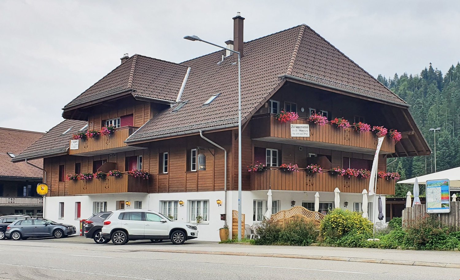 A multi-story wooden building with a tiled roof, balconies, and flower boxes. There are cars parked in front of the building.