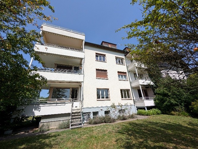 White building with balconies on all sides, garden, many windows, staircase, shrubs and plants.
