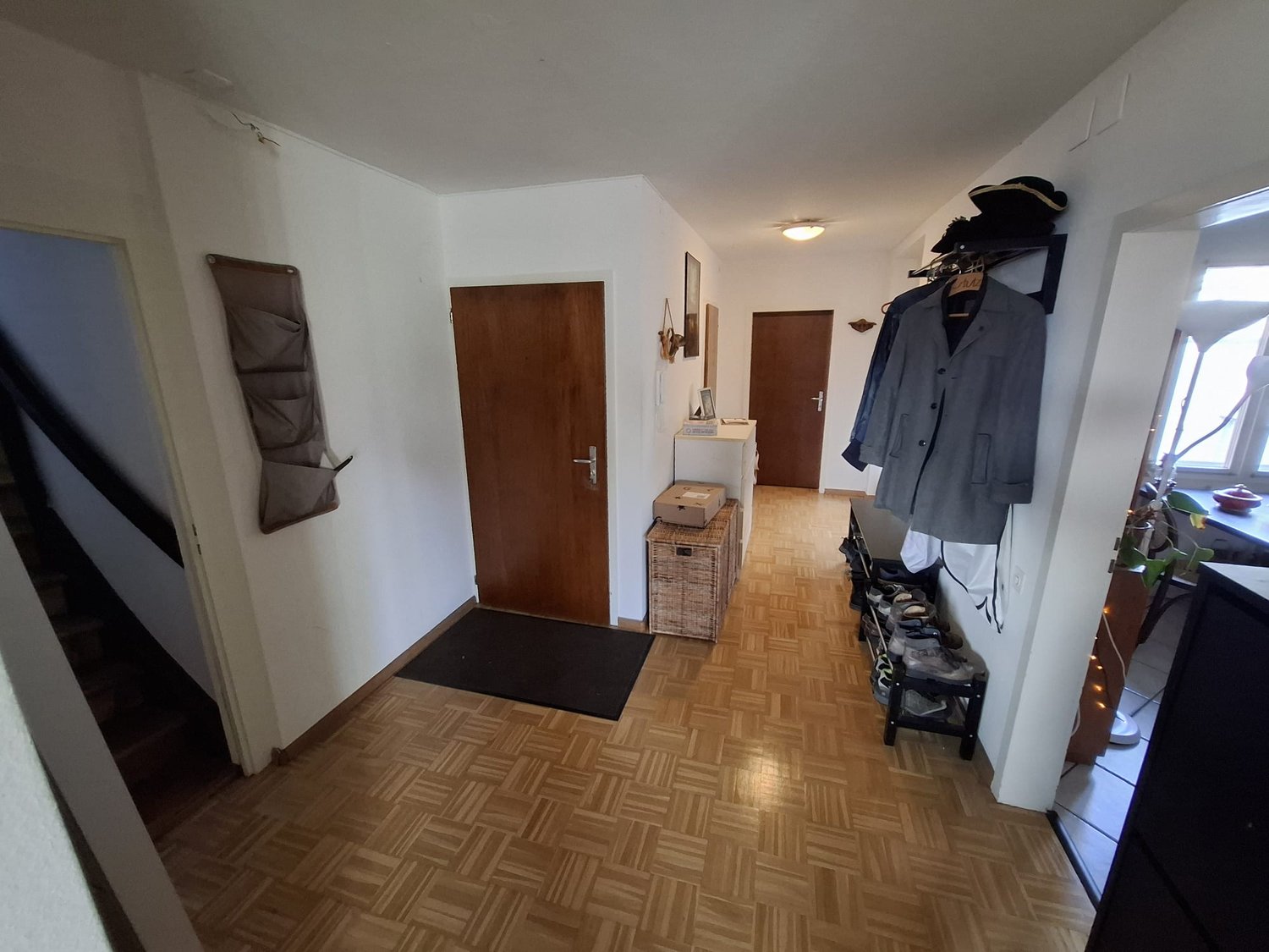 wooden floor, two doors, black mat, rack with hats, clothing, basket, table with box, potted plant