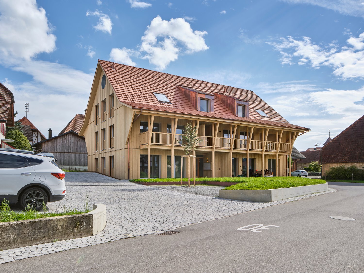 Large two-story wooden house, red roof, wooden balconies, modern design, large windows, front yard, gravel driveway, parked car