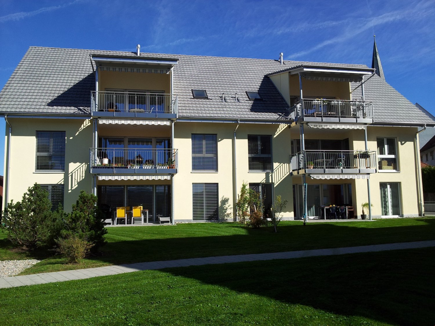 Three story apartment building with three balconies, gray roof, glass doors, windows with shutters, greenery