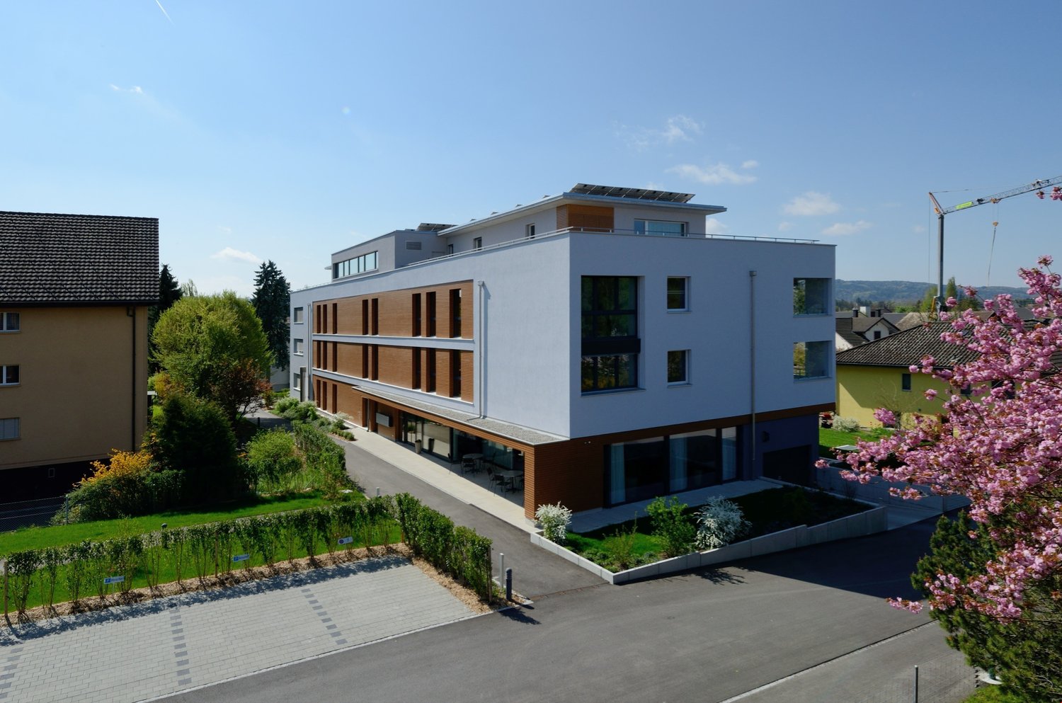 White building, 2 stories, solar panels on the roof, brown panels on the first floor, driveway, trees, green landscape, pink cherry blossoms