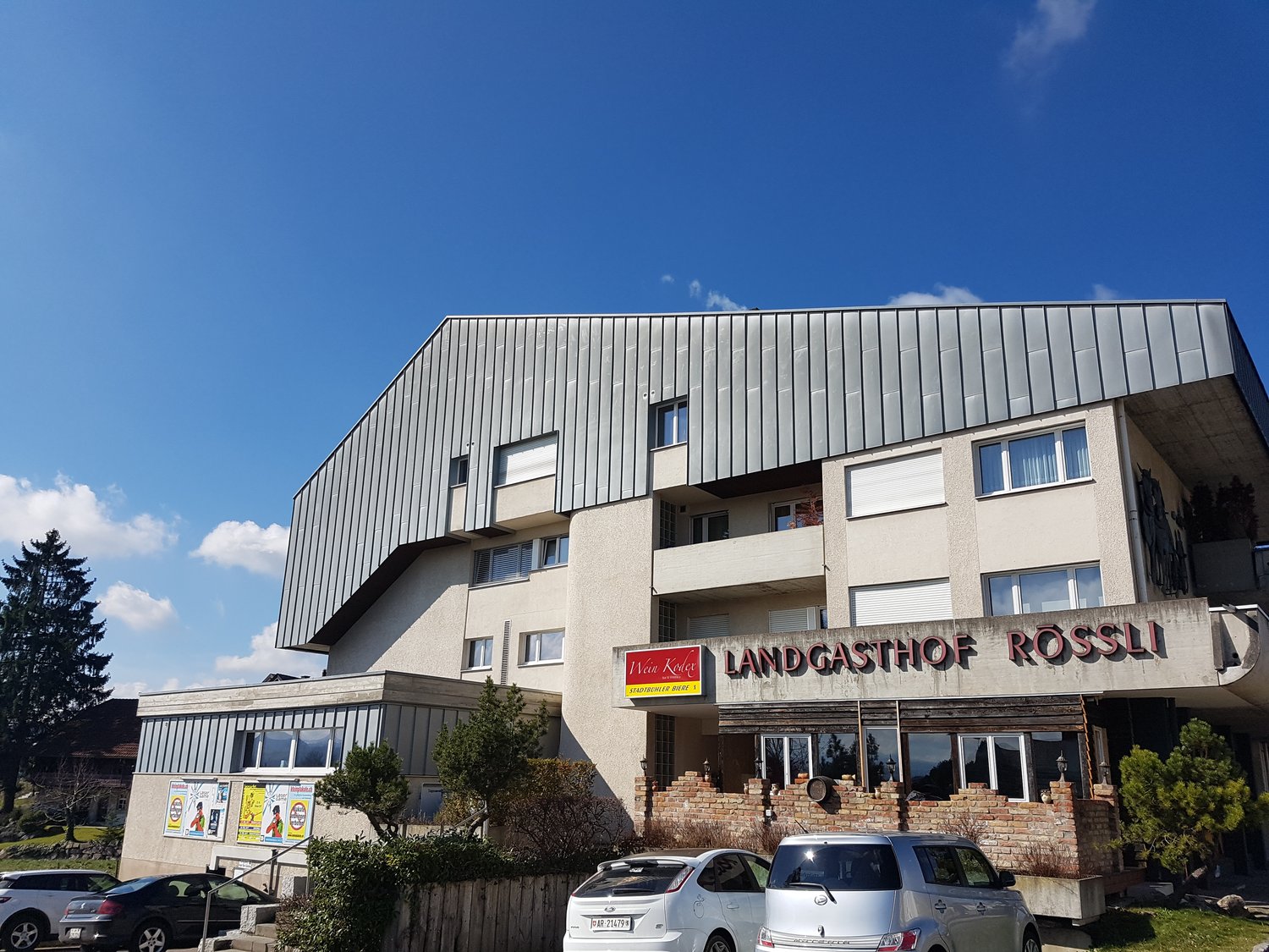 Modern apartment building, large windows, grey roof, red sign with name 'LANDGASTHOF ROSSLI', parked cars in front, brick walls at entrance, trees around the building