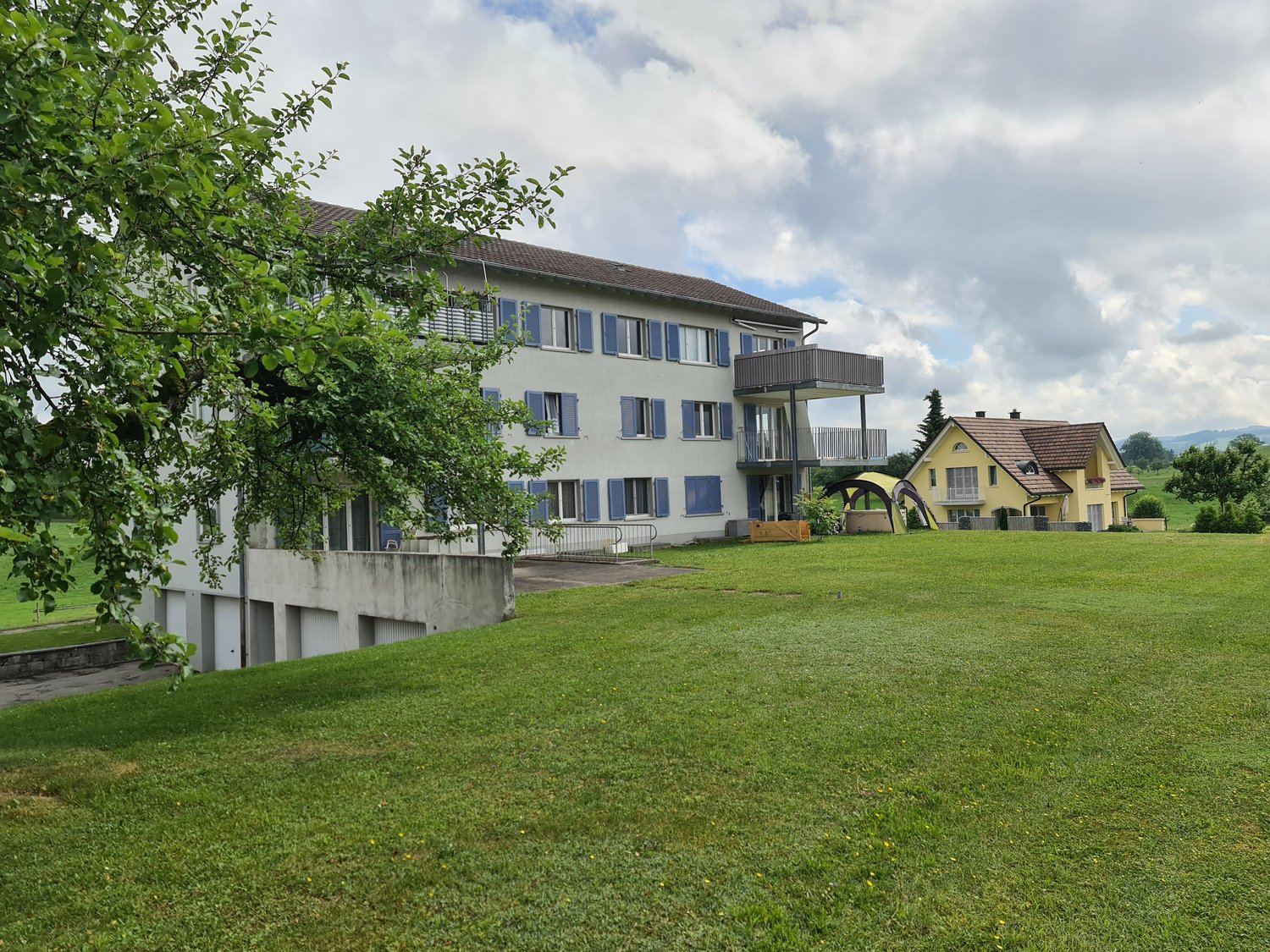 Two story white building, blue shutters, balcony on second floor, grass field in front.