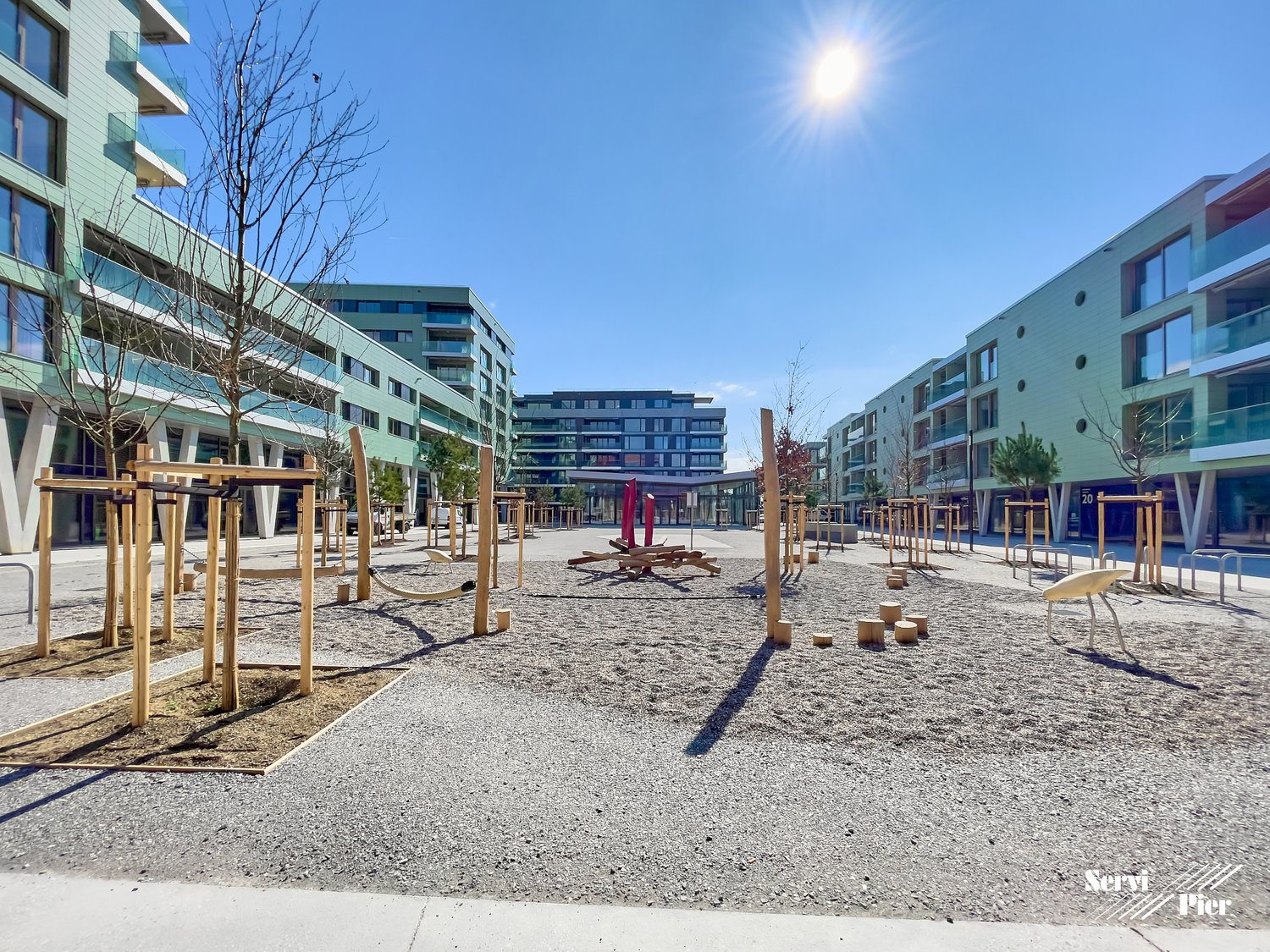 The image shows a public plaza or outdoor space in an urban setting. The area is paved with gravel and features various wooden structures, possibly seating or play equipment. The surrounding buildings have balconies and appear to be modern, multi-story re