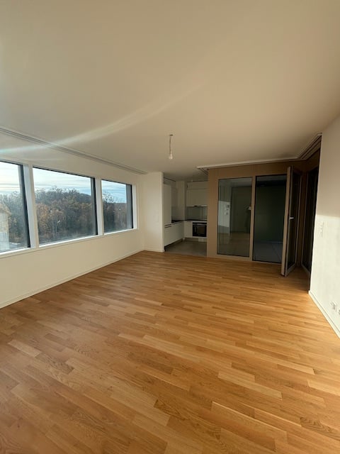 Empty room, hardwood floors, white walls, large windows with view, kitchen and balcony visible