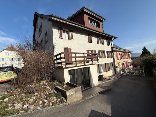 Two-story building, white walls, brown roof, windows with shutters, balcony, pathway in front, cars parked on the side