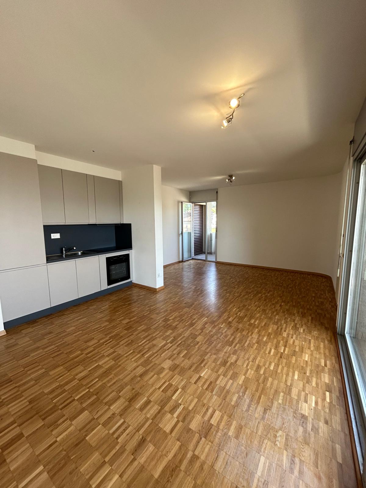 The image shows a spacious open-plan apartment with a modern kitchen and living area. The apartment features hardwood flooring throughout, white walls, and recessed lighting. There is a sliding glass door leading to what appears to be a balcony or terrace