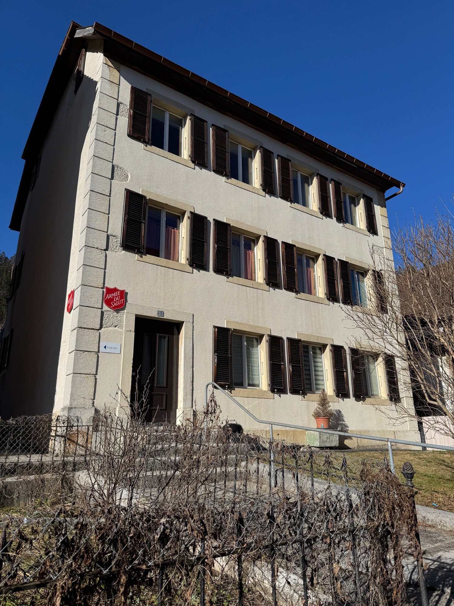 3 story apartment building, beige exterior, multiple windows, dark brown doors, stairs with railings, green shrubbery, sign