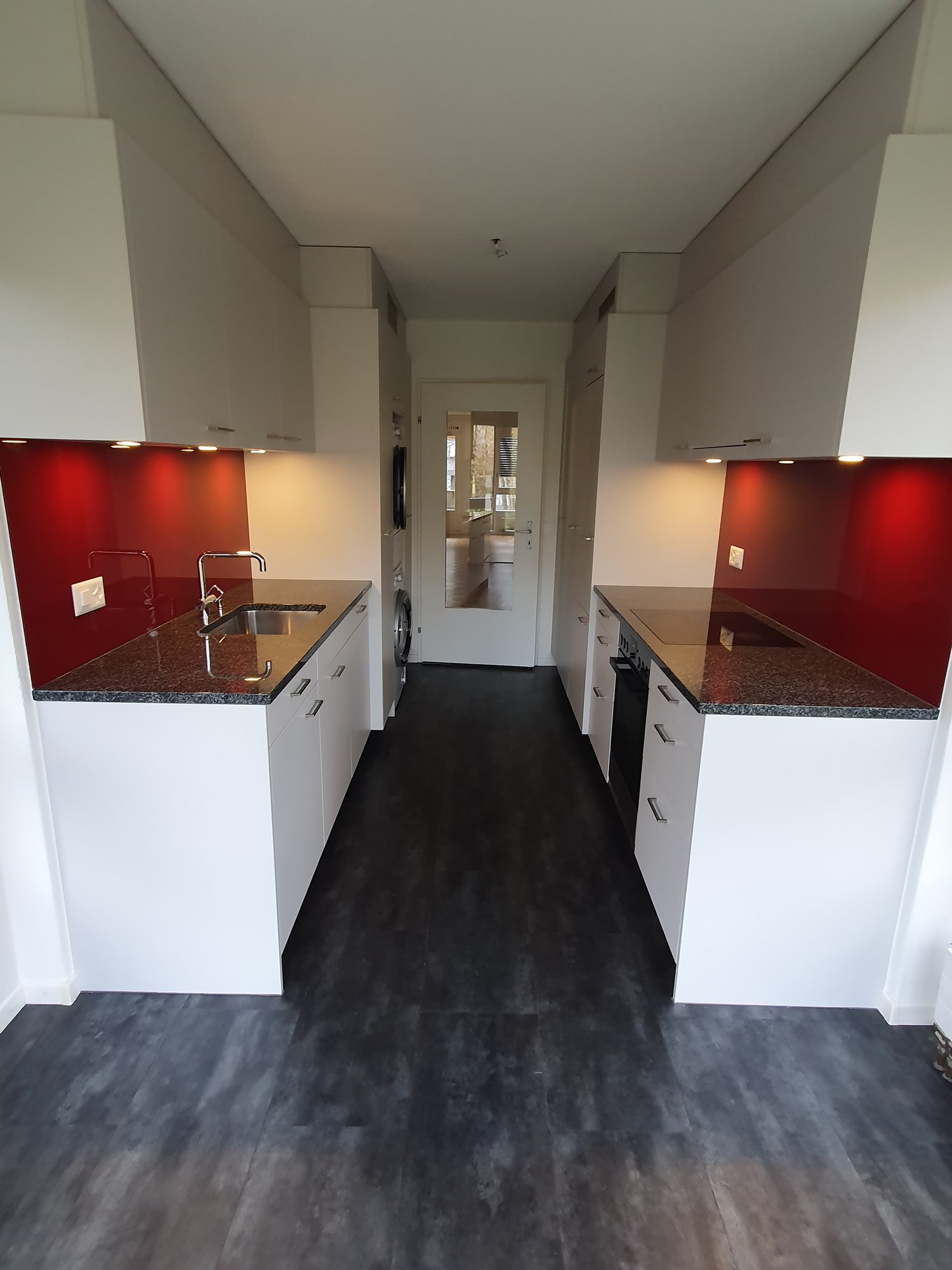 modern kitchen with white cabinets, black countertops, red backsplash, stainless steel sink, dishwasher, washing machine, black floors, pendant lights