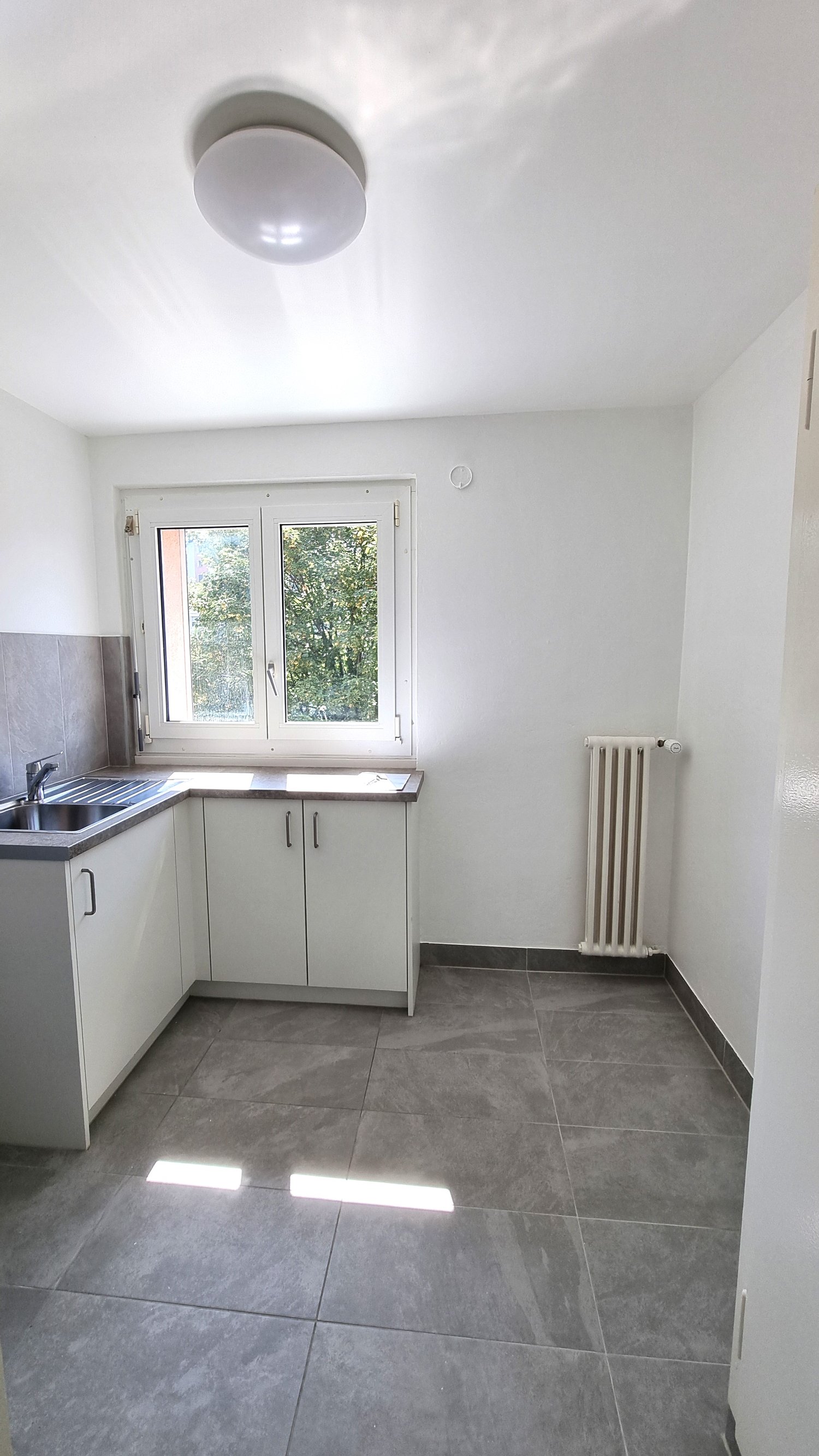 White cabinets, gray tiled floor, window with radiator, stainless steel sink, white ceiling light