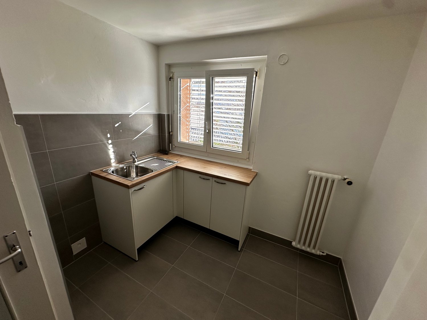 an empty kitchenette, stainless steel sink, cabinet, tile flooring, white radiator, small window
