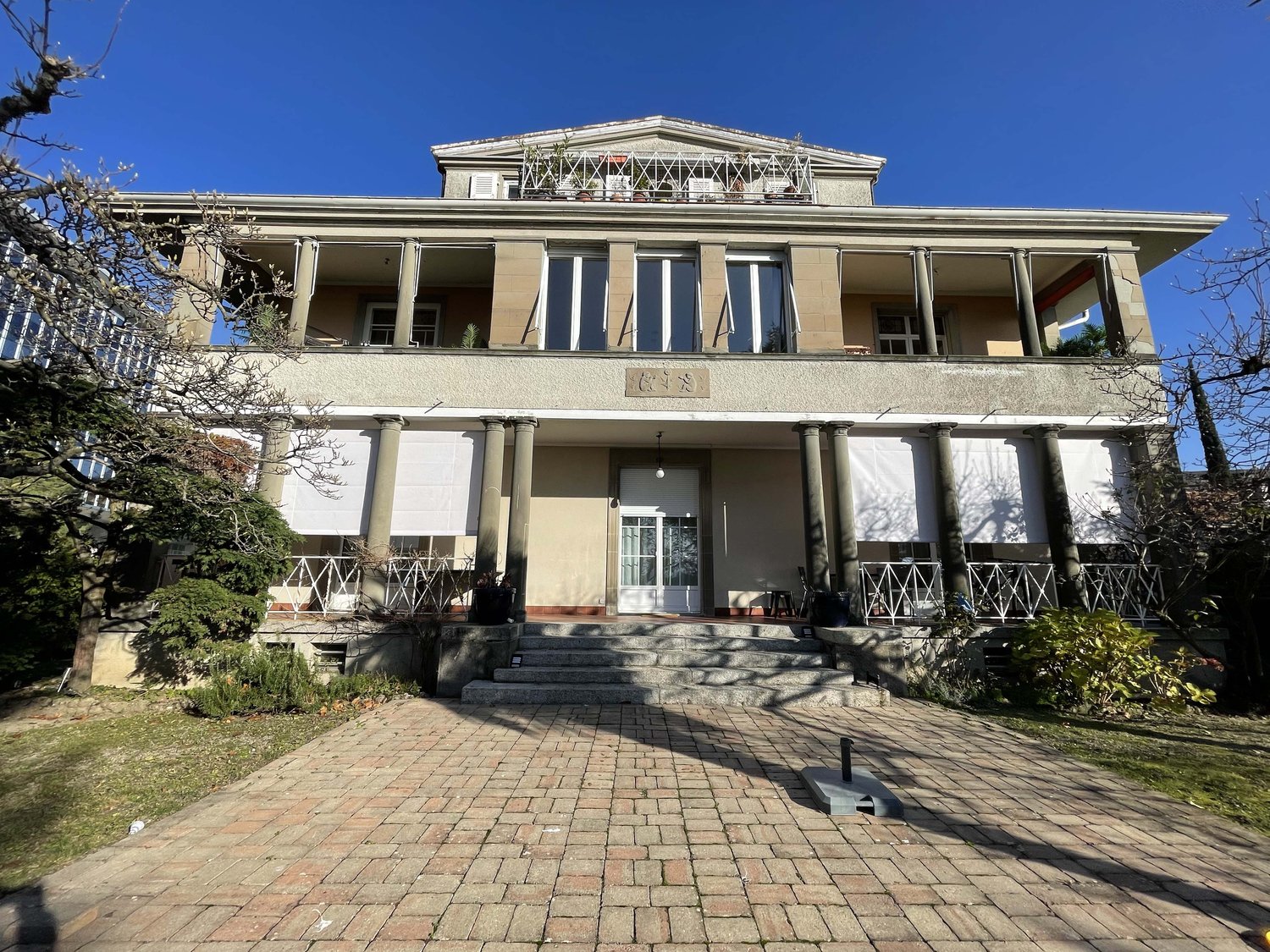 2 story building, stone and concrete facade, large windows, terracotta tiled front, brick pathway, stairs leading up to entrance, terrace with metal railings