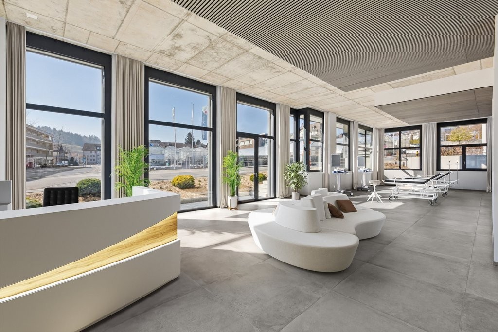 Large open-plan office space with floor-to-ceiling windows offering a view of the outdoors. The space features a modern, minimalist design with white furniture, including curved sofas and chairs. The floor is covered in gray tiles, and the ceiling has a g