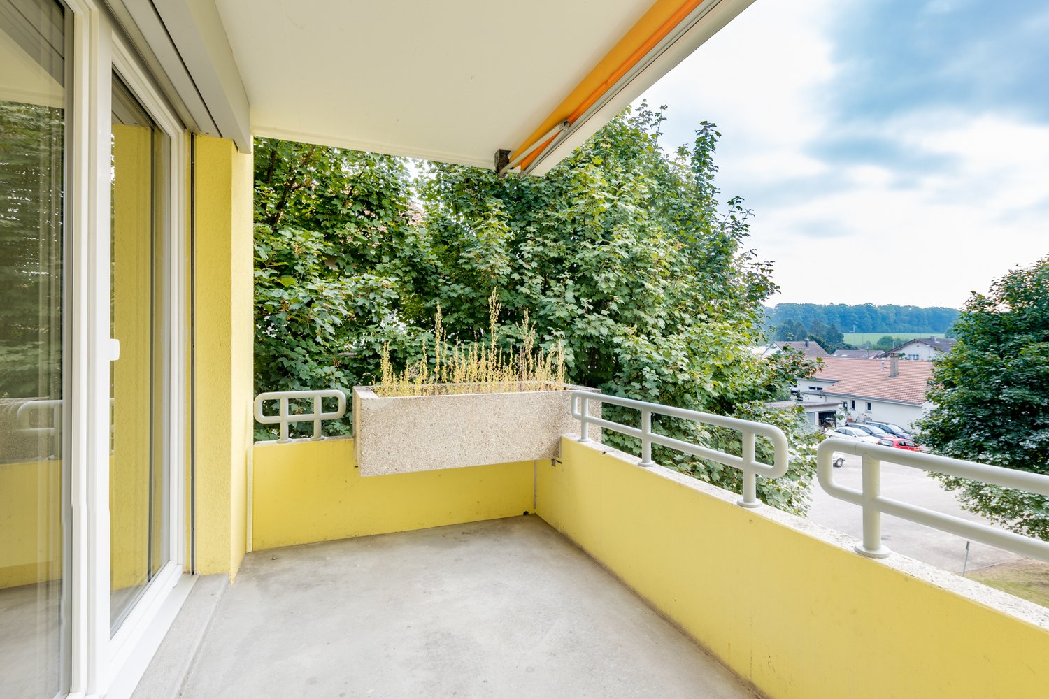 Yellow painted walls, concrete balcony floor, white railing, a plant box, open glass door to the interior