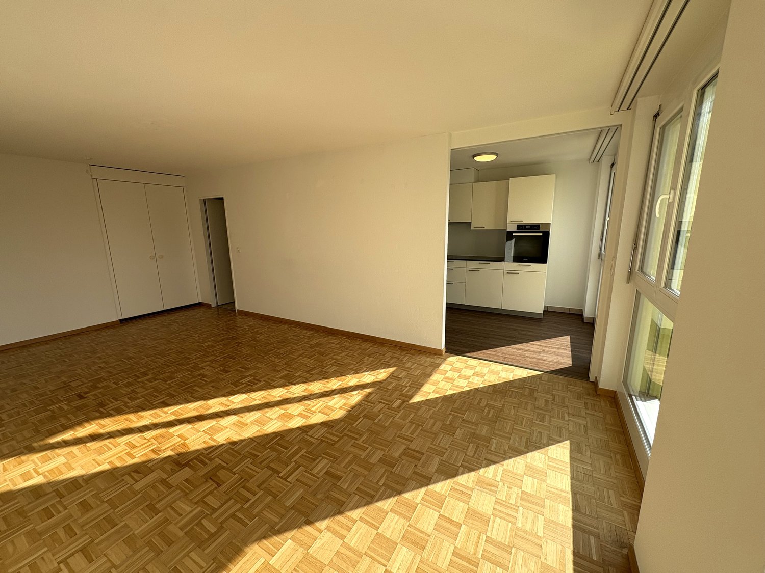 Wooden flooring, sun-drenched, large windows, empty room, white kitchen with black oven, white cabinets.