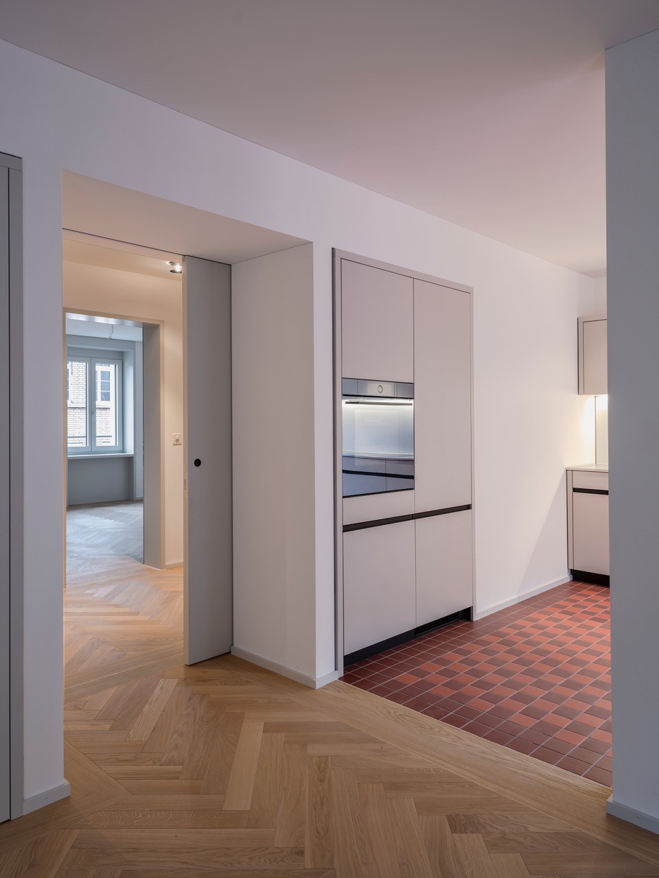 Minimalist modern kitchen, white cabinets, wooden floor, red tiles near oven, sliding door leading to another room, open window in other room.