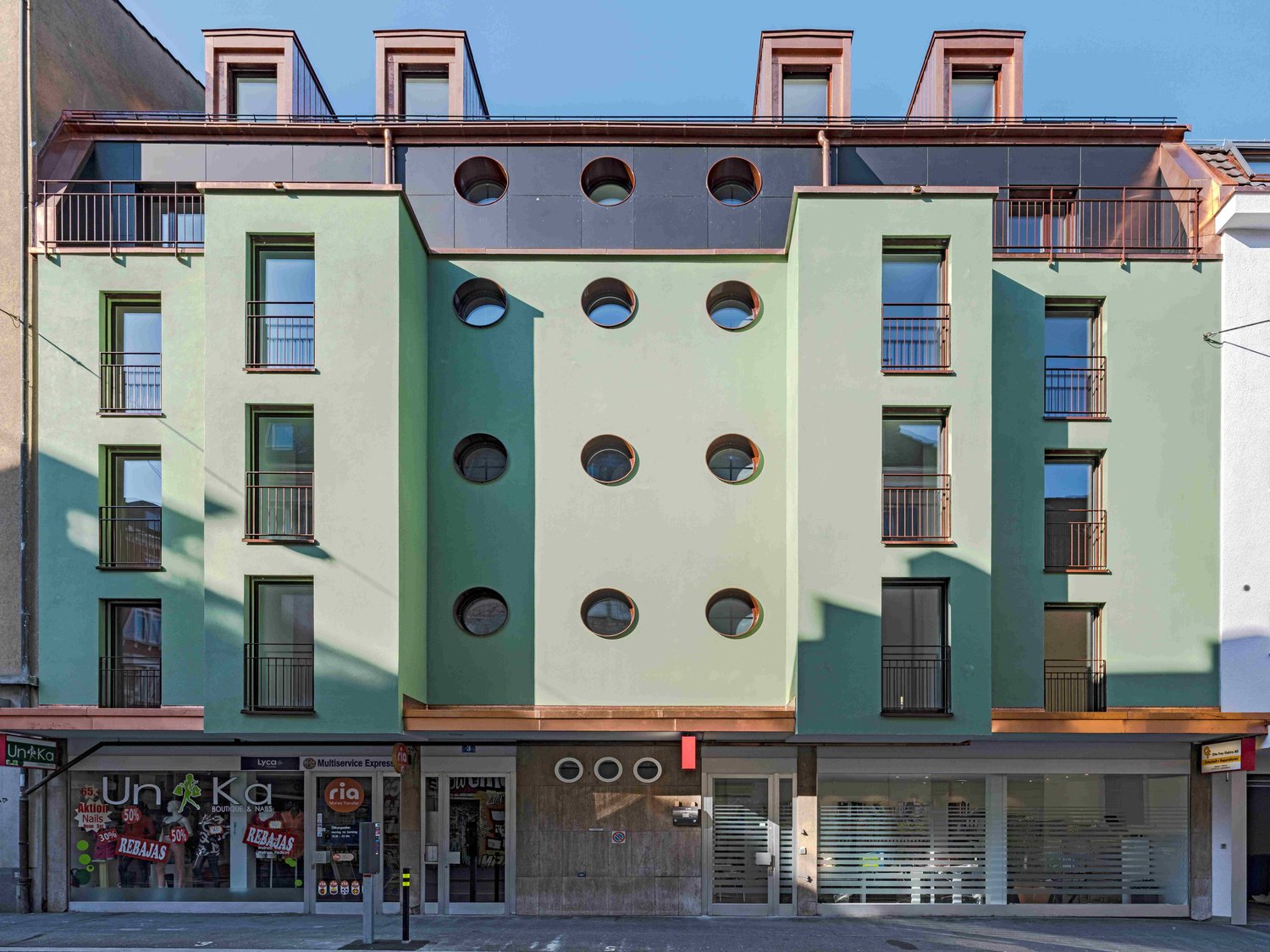 A multi-story commercial building with a unique architectural design. The building features a green and gray color scheme, with circular windows and balconies. The ground floor has several retail stores, including a store called 'Unka'.
