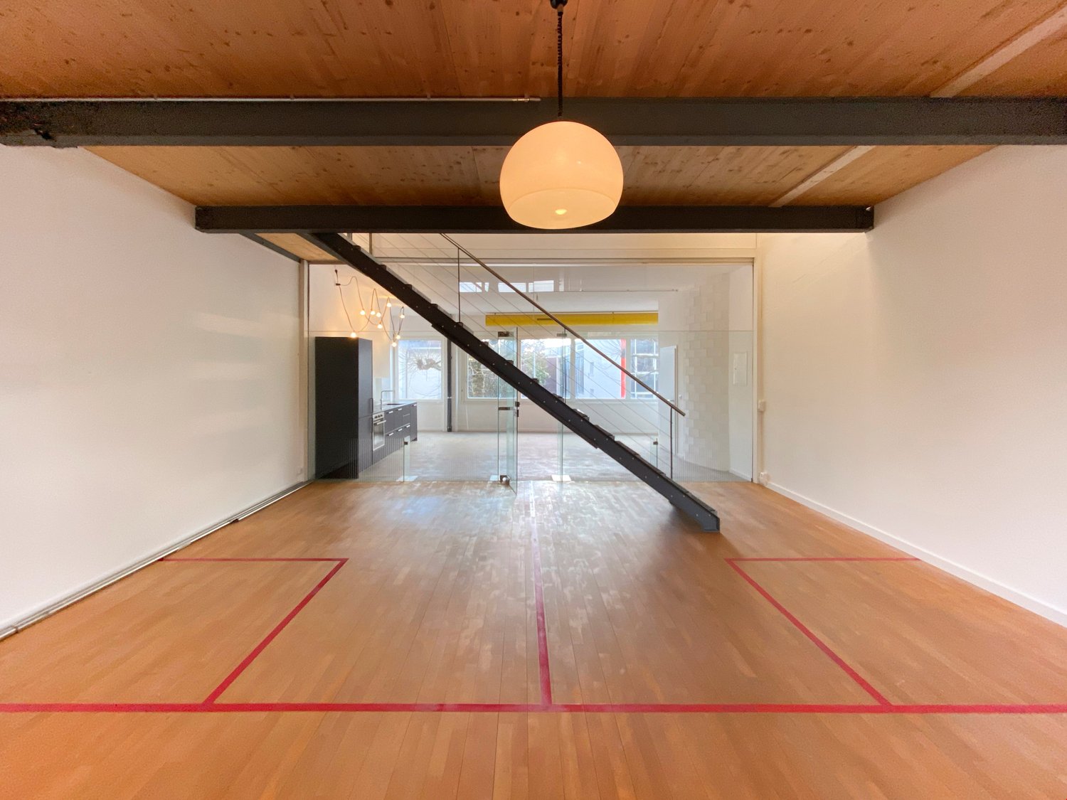 Open space with wooden floor, white walls, glass partition, staircase, hanging lamp, kitchen area, and storage unit.