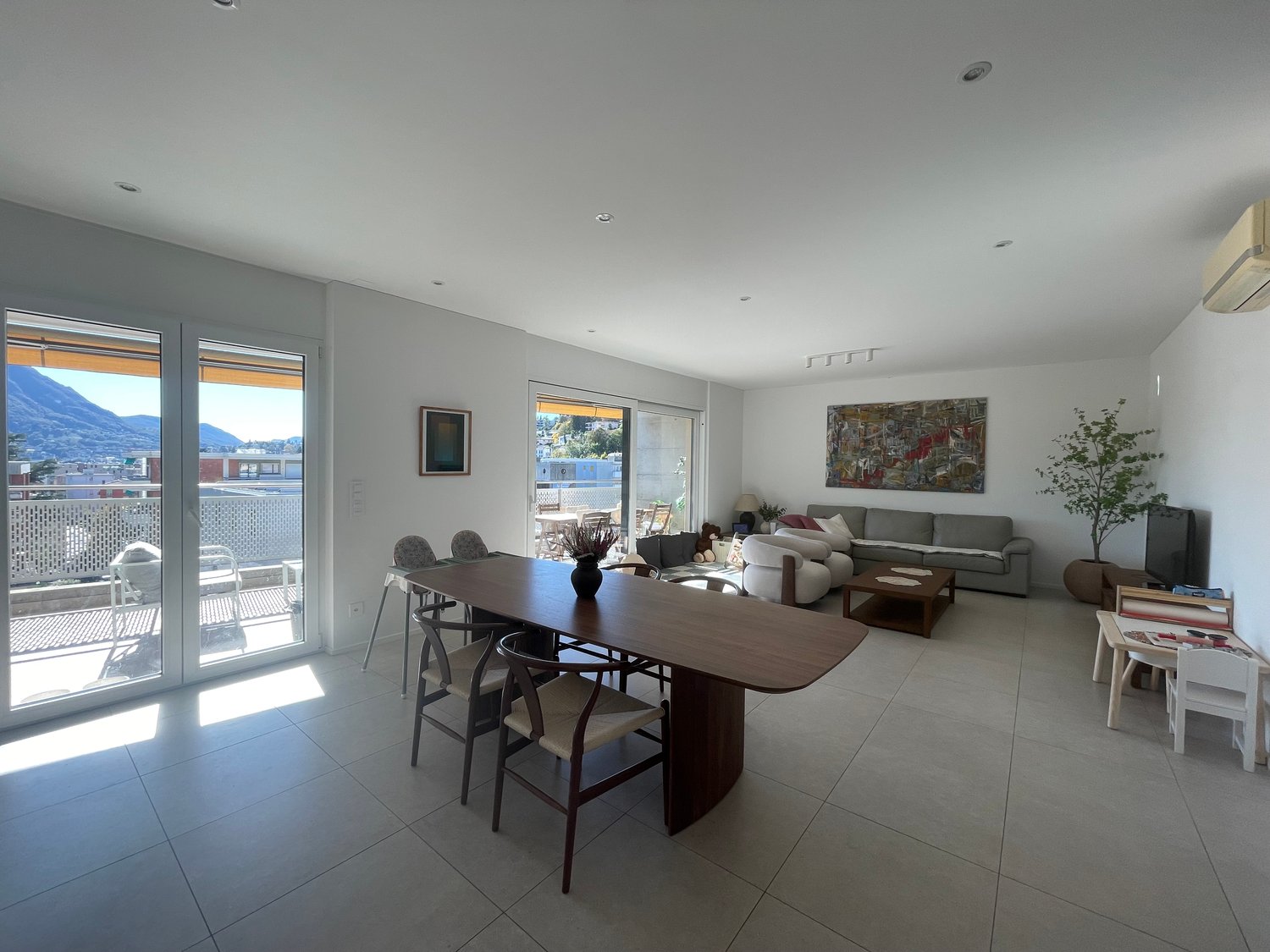 Modern dining room with large glass doors leading to a balcony, white tile floor, gray sofa, dining table, paintings on the wall, indoor plants