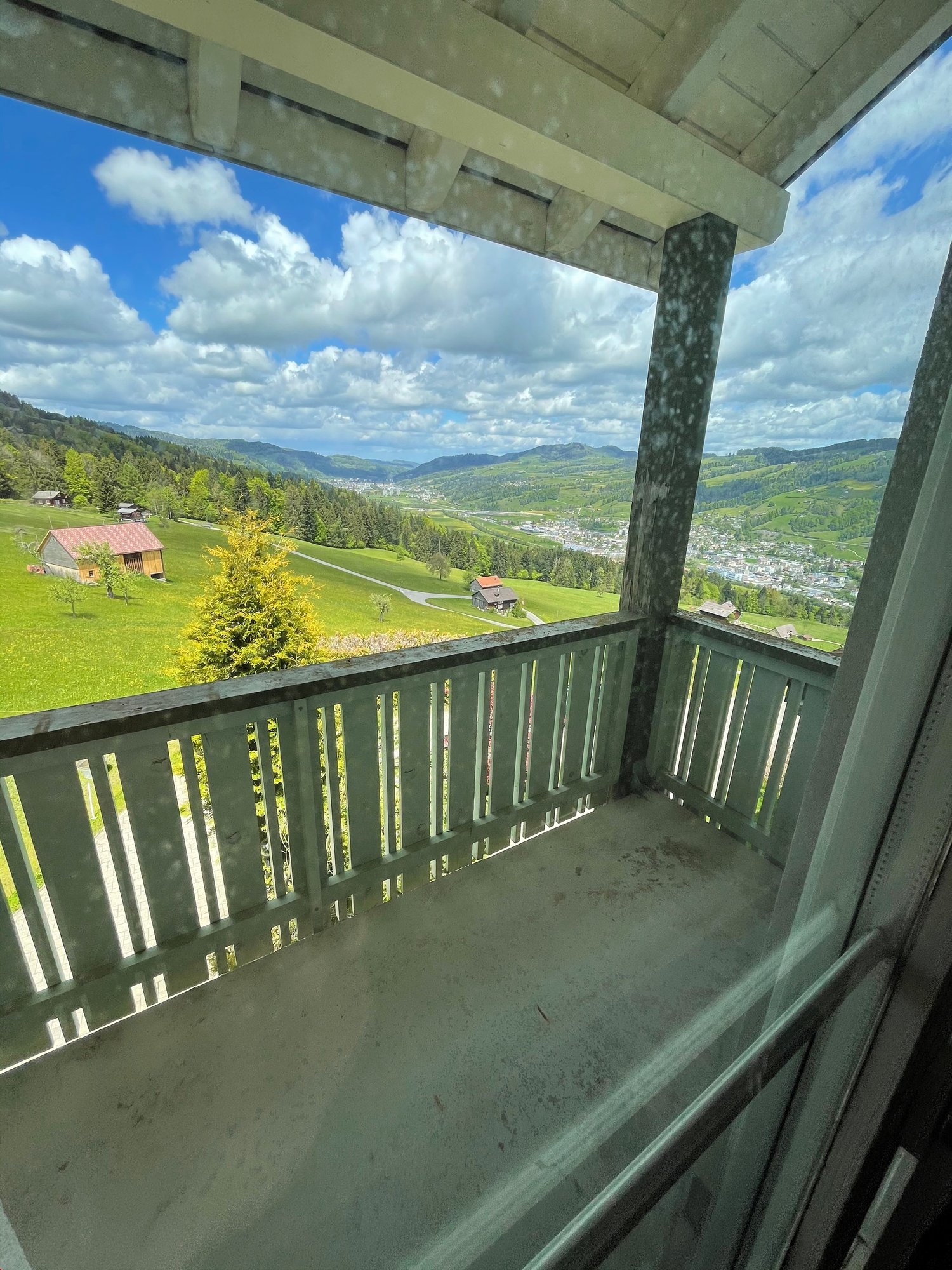 Balcony with a scenic view of the countryside, including rolling hills, forests, and a small village in the distance. The balcony has a wooden railing and overlooks the lush, green landscape.