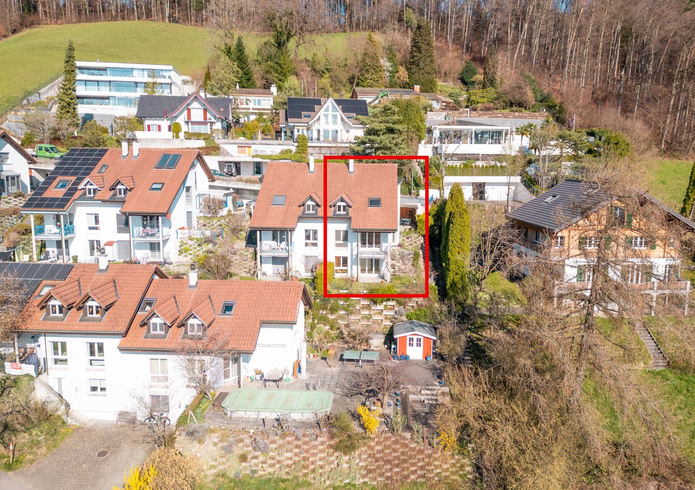 Aerial view of a residential area with several houses, one of which is highlighted with a red square. The highlighted house has a brown roof and white walls. The houses are surrounded by trees and greenery. The area is on a slope, with some houses having