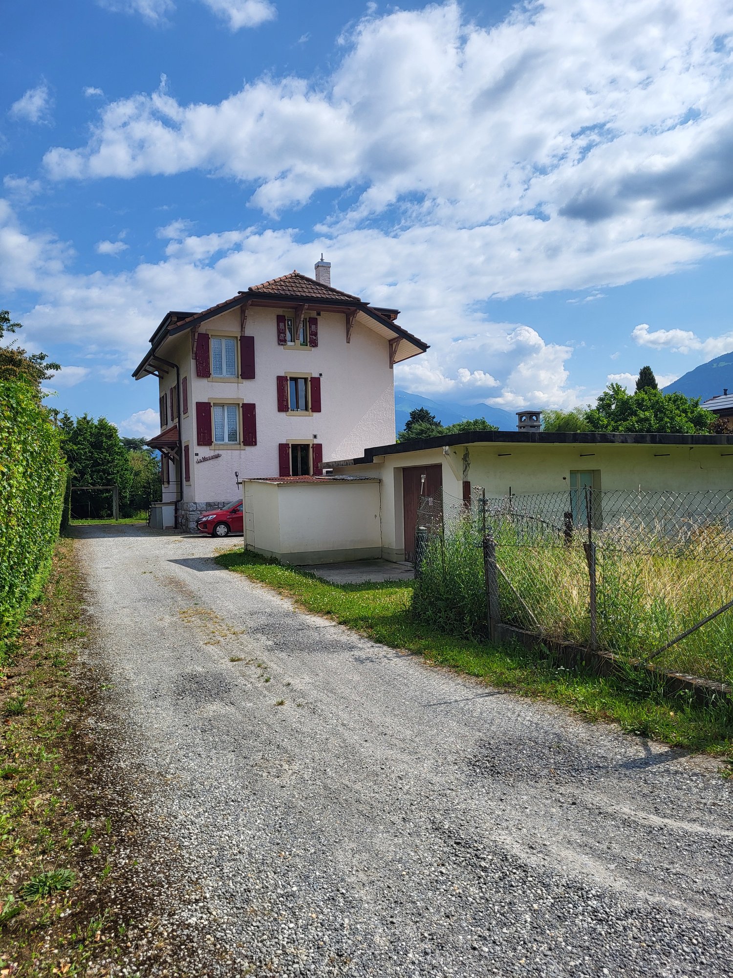 Two-story house, red car parked, gravel driveway, garden, mountains in the background