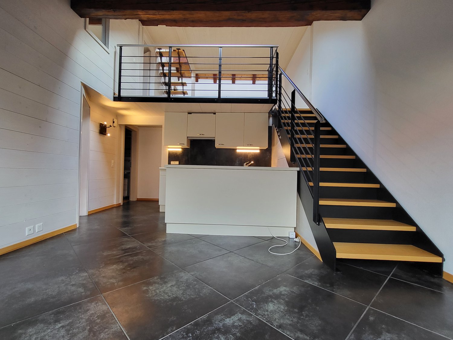 Black tiled floor, white walls, wooden railing, open kitchen with cabinets and a sink, black and yellow stairs, an upper floor balcony, and wall-mounted lighting