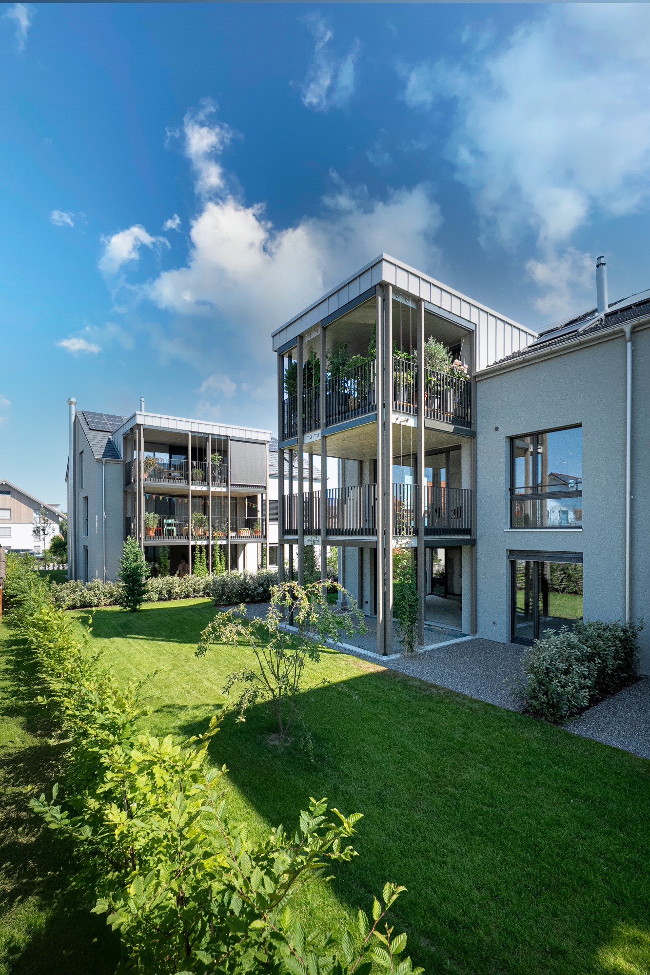 Multi-story apartment building with balconies, surrounded by a well-maintained garden and lawn area, with parking spaces available.