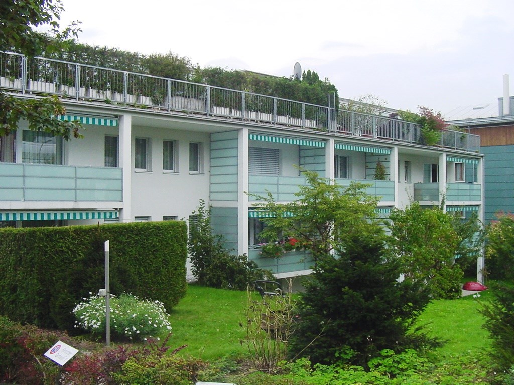 Multi-story apartment building with green and white exterior, balconies, and surrounded by lush greenery and landscaping