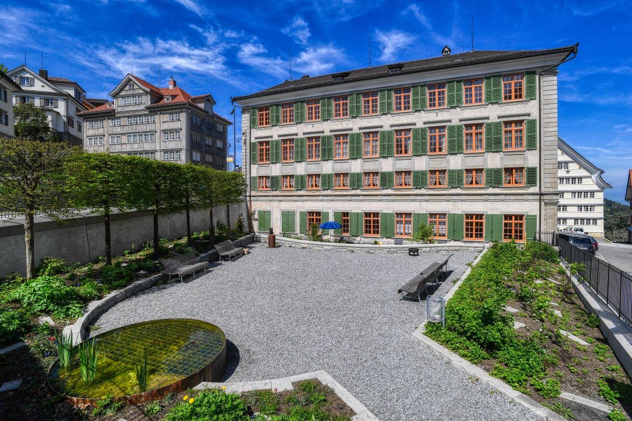 Large building with gray walls and red roof, large windows with green shutters, central courtyard with gravel, circular pond, small shrubs, wooden benches, black fence