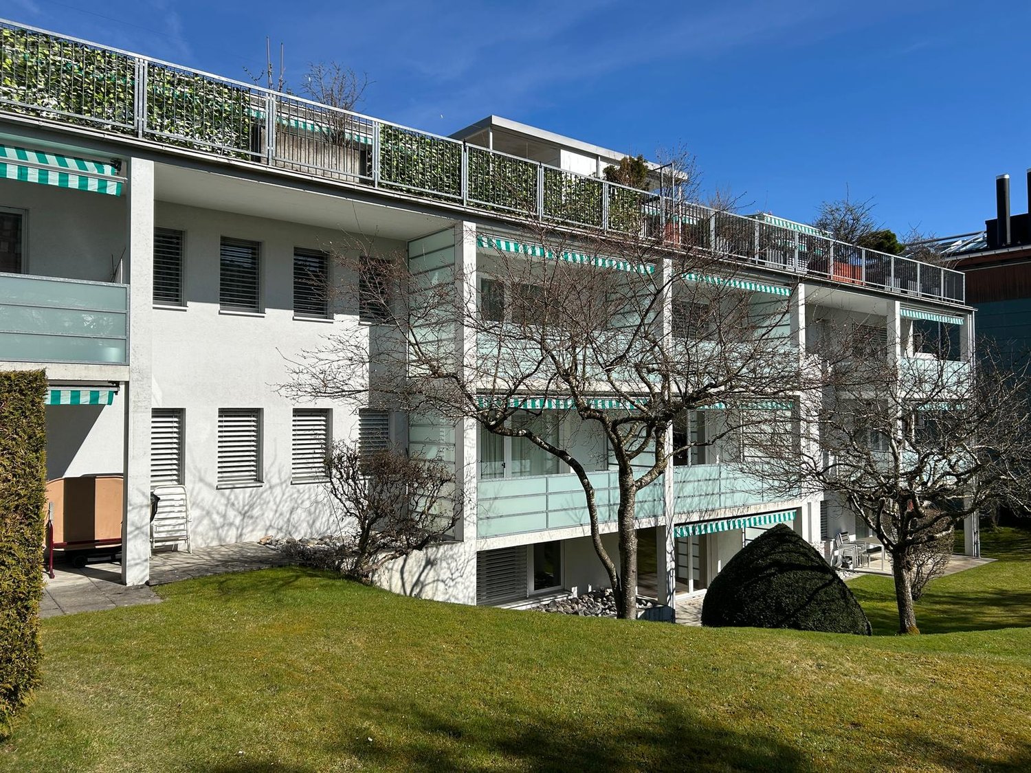 White building, balconies, grass, trees, shrubs, paved walkway, greenery on top of building