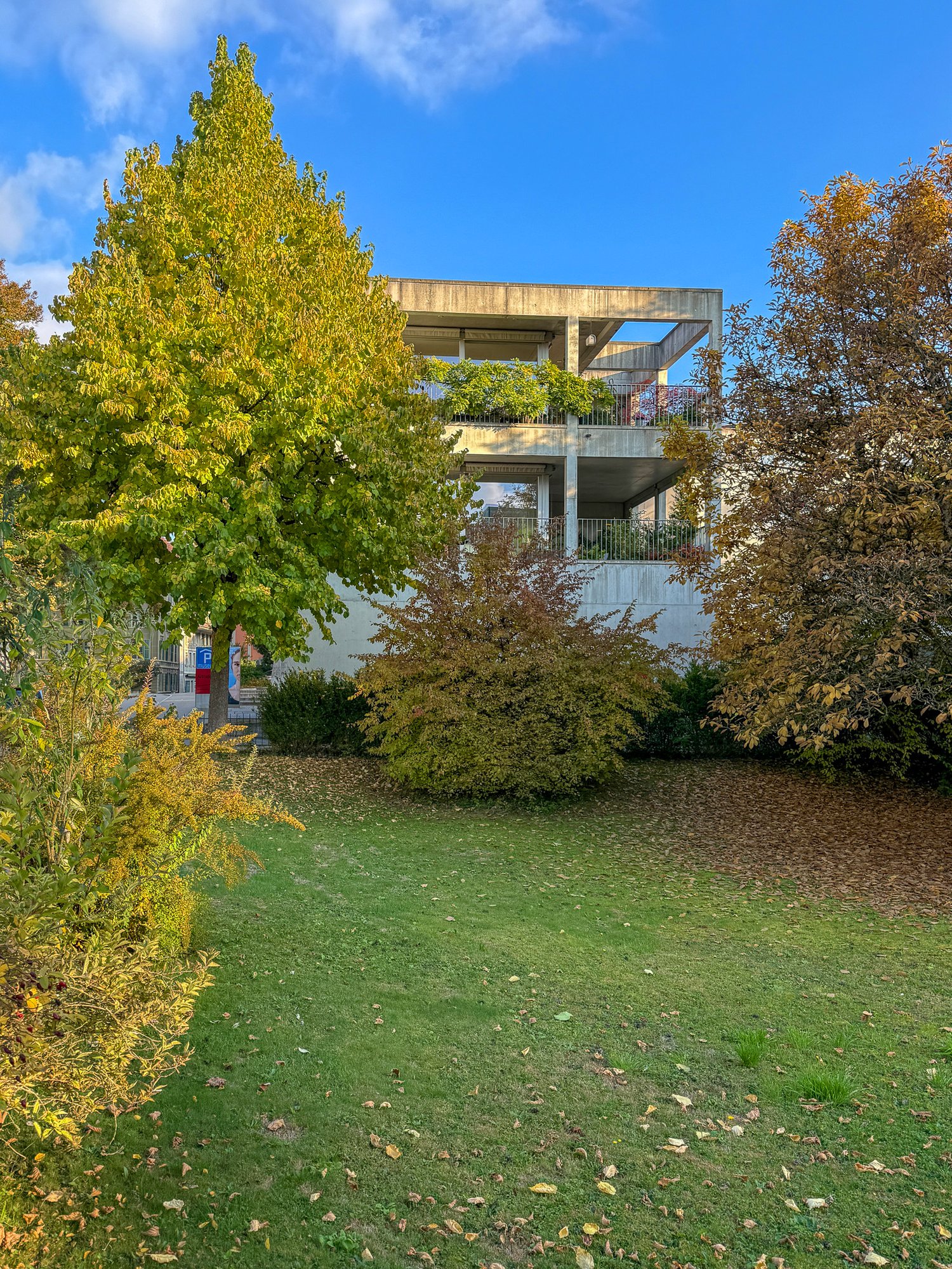 Concrete building with raised ground floor, balconies on upper level, surrounded by grass and trees.